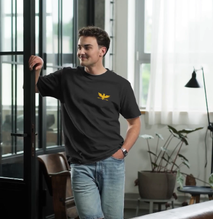 A man wearing a black crewneck t-shirt with a yellow eagle logo on the chest, paired with light blue jeans, standing in a modern room with large windows.