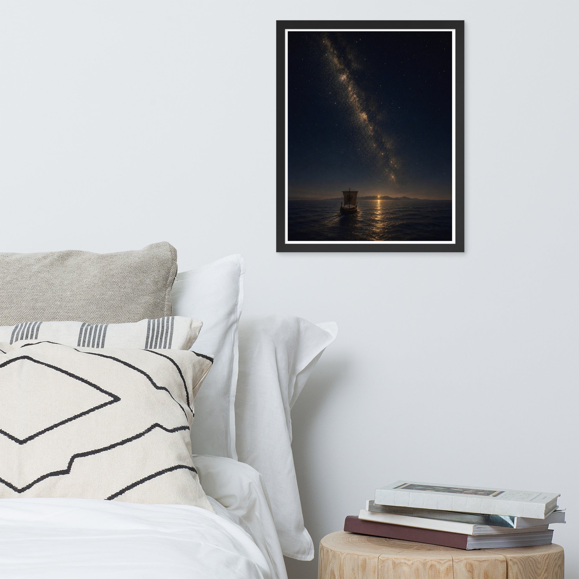 A framed wall art print depicting a night ocean scene with the Milky Way over a lighthouse, styled above a bed with pillows and next to a small wooden side table with stacked books.